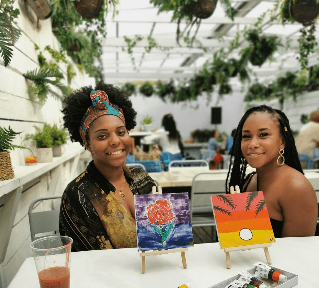 Two women sit at a table in front of mini painted canvases, smiling