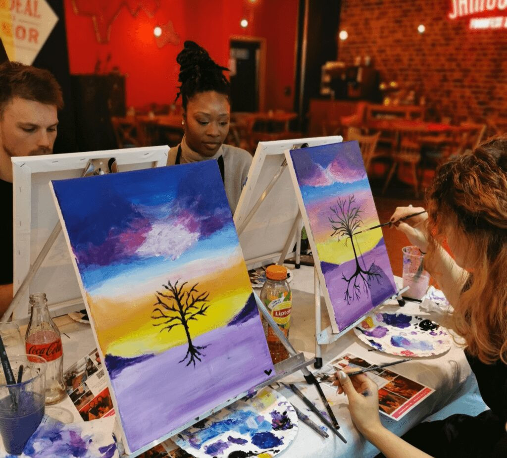 A group of people sit at easels at a table, painting colourful landscape scenes