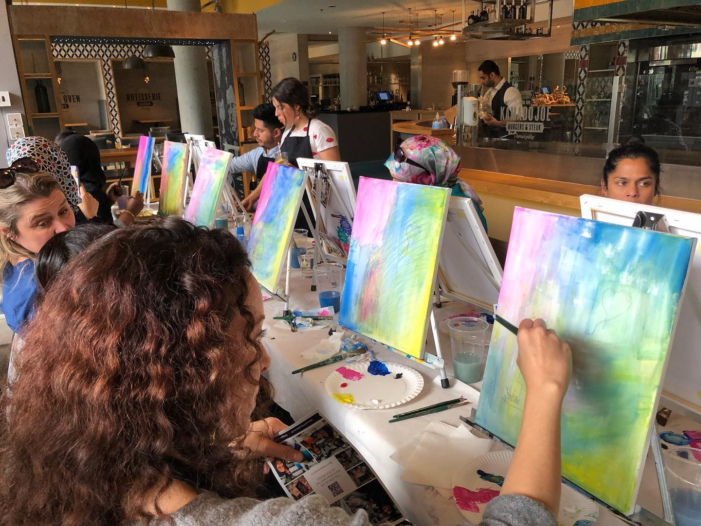 A group of people sitting at canvases at a table, painting colourful pictures at a painting class
