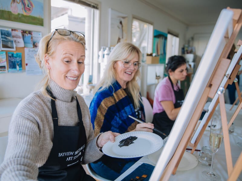 Three women are sitting at easels, painting at a sip and paint class