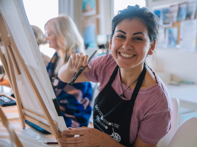 A smiling woman sitting at an easel at a paint and sip experience day for her
