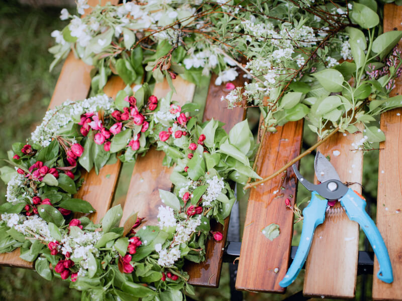 A floral wreath made from pink and white flowers and green foliage lies on a table next to some secateurs