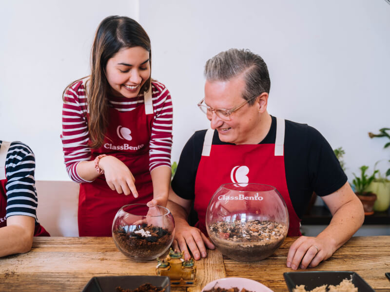 A man and woman are making a glass terrarium at a wooden table