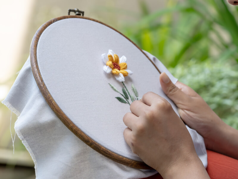 A close up of a person embroidering fabric in a hoop with a flower and leaves in embroidery silks