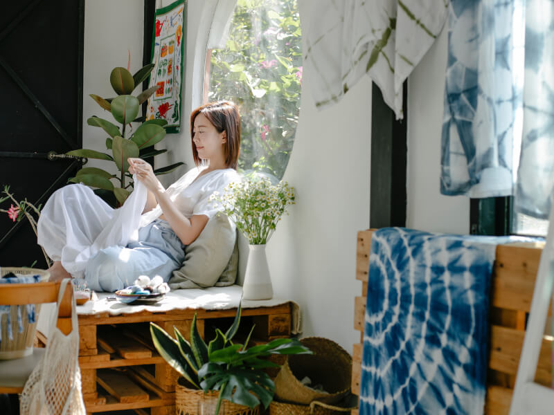 A woman sits cross legged in a bright room, sewing white fabric by hand