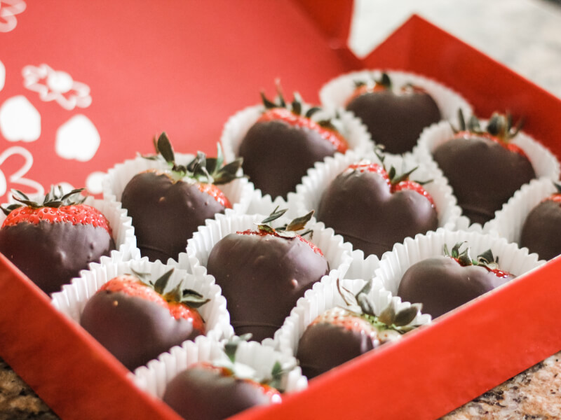 An open red cardboard box displays a selection of strawberries in white paper cases dipped in milk chocolate