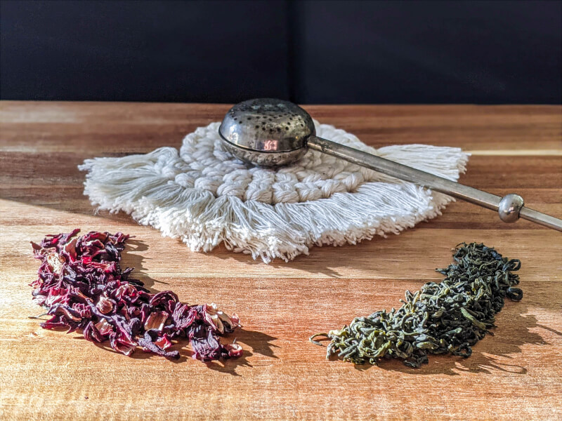A macrame coaster lies on a wooden table, with a tea strainer on top and loose leaf tea surrounding it