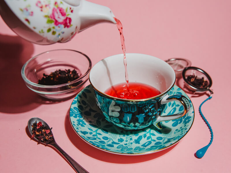 A close up of a person pouring pink tea from a colourful teapot into a teacup