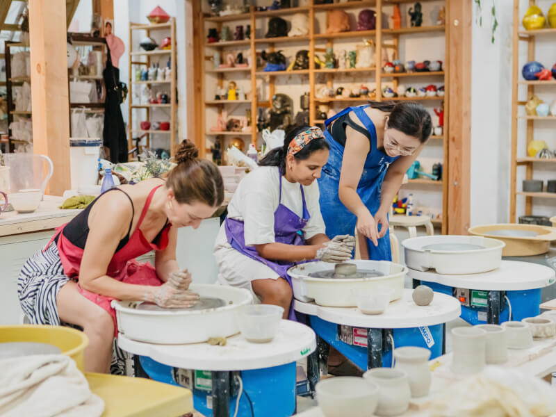 Two women are using pottery wheels to make handmade clay items, guided by a third woman who is standing nearby