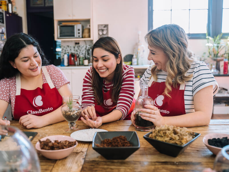 Three women are sitting at a wooden table, making glass terrariums 