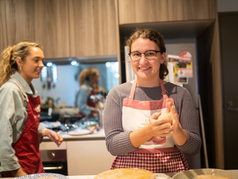 A woman in an apron and a grey jumper is rolling dough between her hands and smiling at the camera