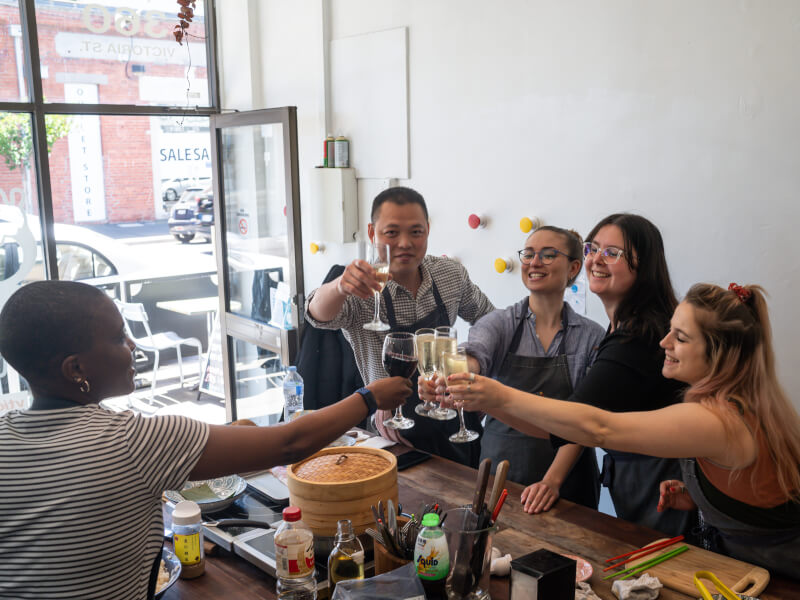 A team of people are sat around a table with cooking accessories, clinking glasses