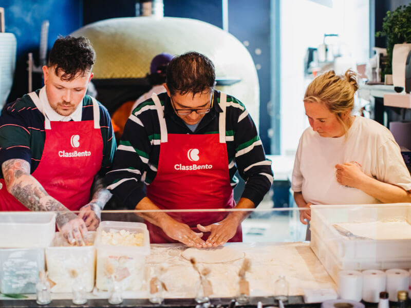 Two men are standing in a kitchen with boxes of ingredients in front of them, adding toppings to homemade pizzas
