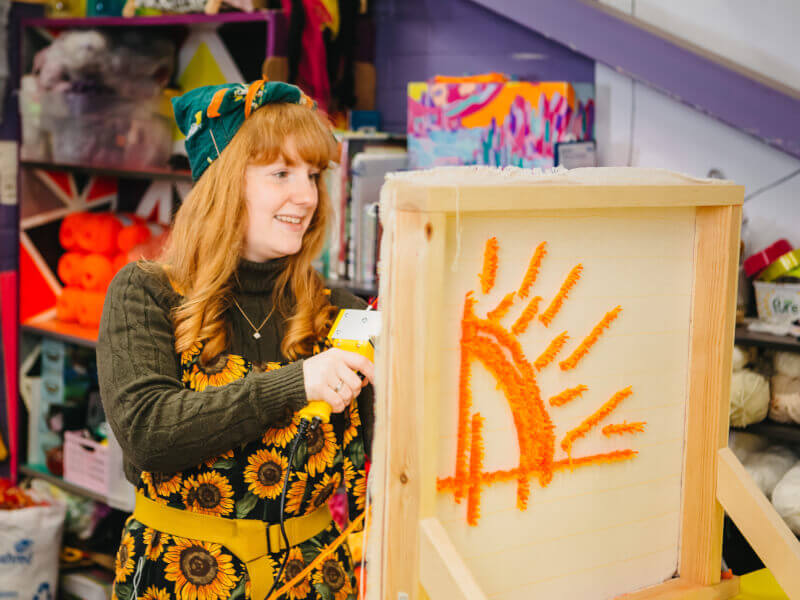 A woman at a needle punching class is creating an orange design on fabric with a tufting gun