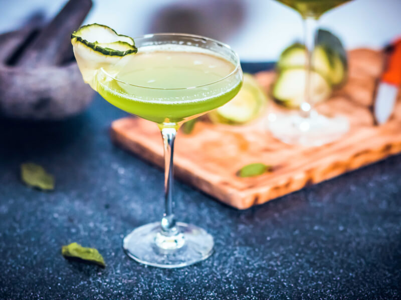 A green cocktail with cucumber curled around the rim of the glass sits on a black worktop. There is pestle and mortar in the background.