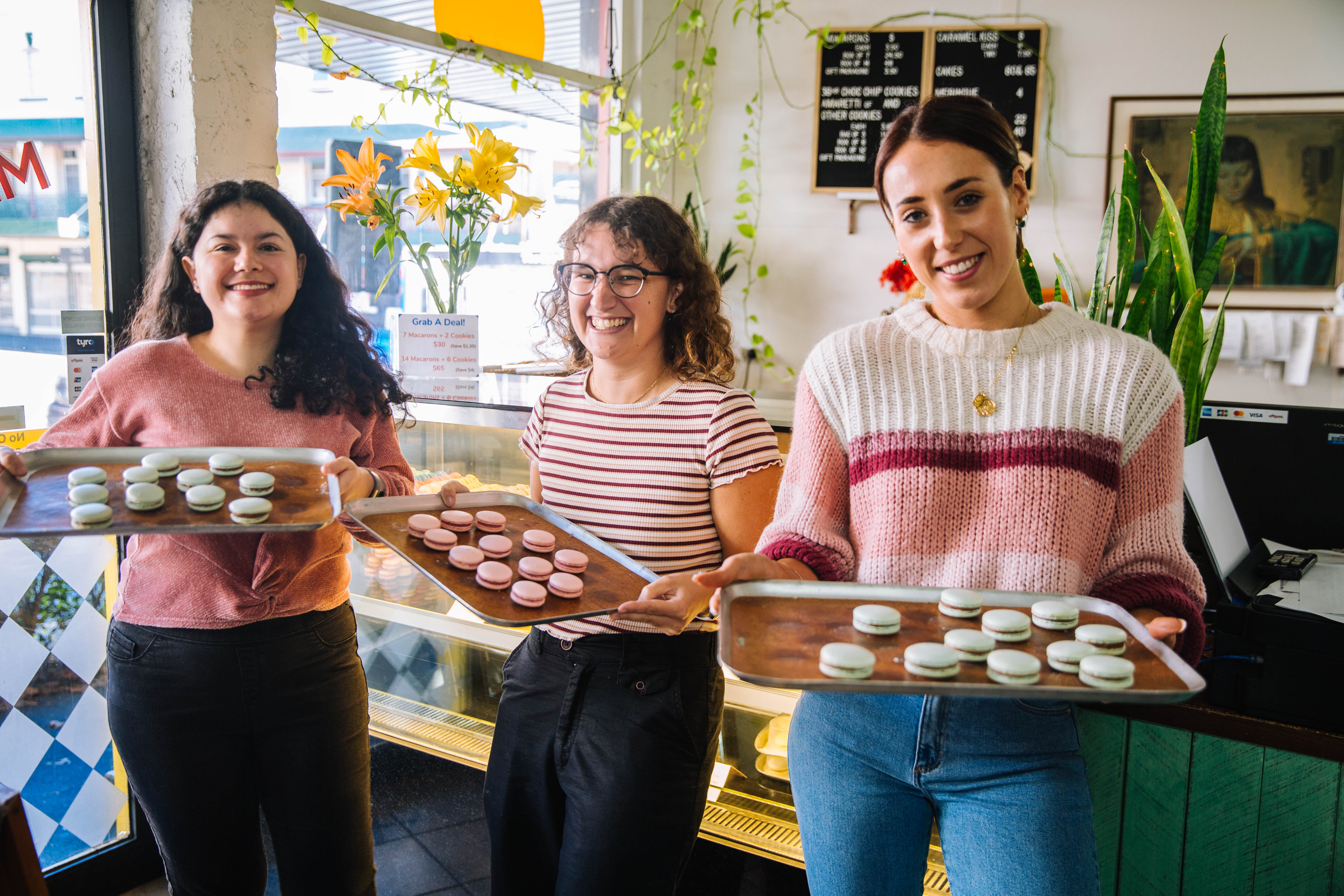 Three women holding trays of colourful handmade macarons