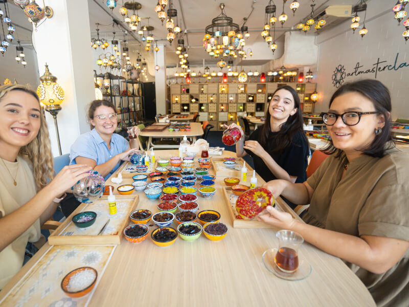 Four smiling women are sitting at a table making mosaic lamps