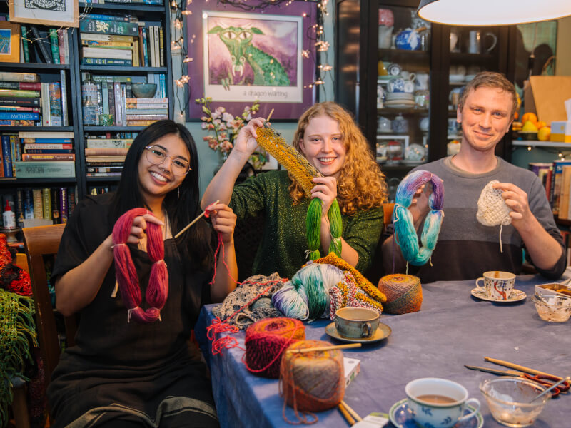Three people are sitting behind a table smiling and holding up yarn and crochet