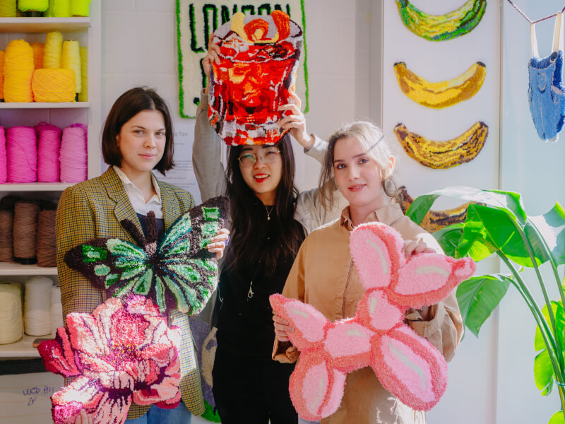 Three women are standing in a studio, holding hand tufted rugs in various colourful designs