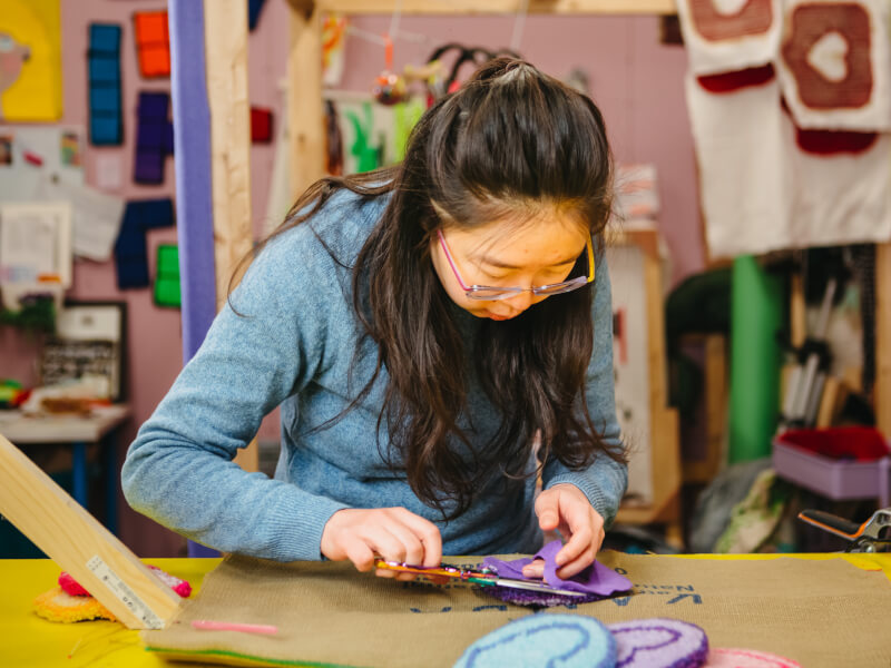 A woman in a blue top is cutting a shape out of purple felt