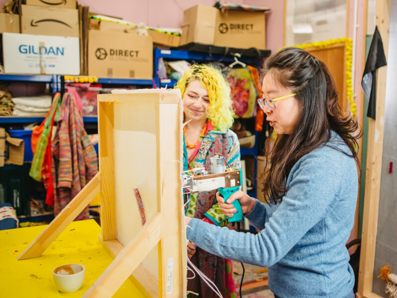 A woman is using a tufting gun to create a design on fabric in pink yarn, while a second woman looks on
