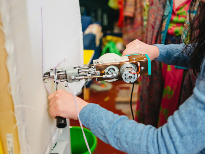 A close up of a woman in a blue top creating a design on canvas using a tufting gun and pink yarn