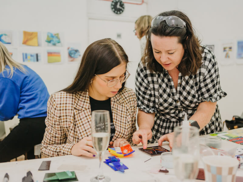 A glass art tutor shows a woman sitting at a table with a glass of wine how to begin a glass art mosaic 