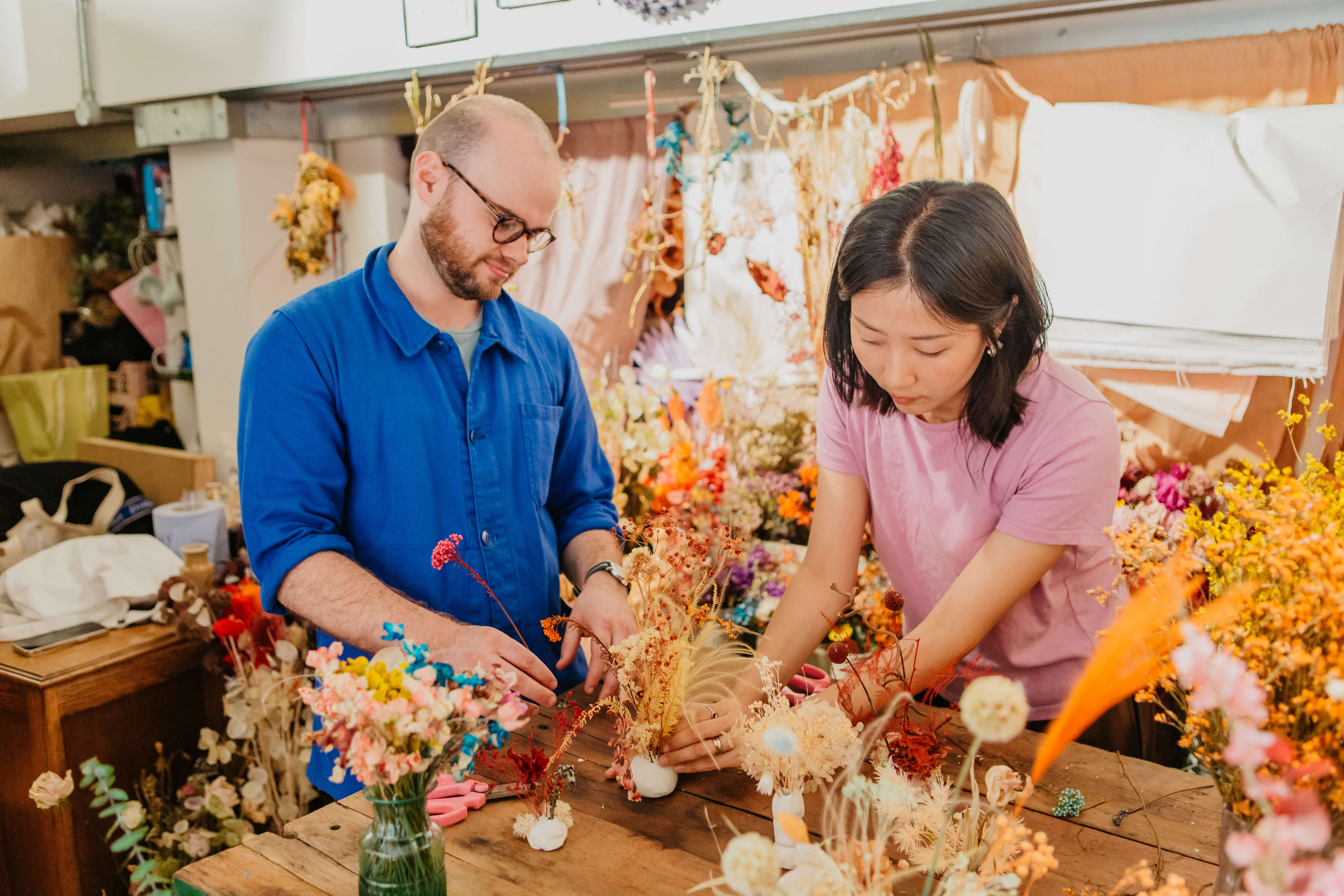 Two people are working on a clay ikebana floral arrangement in a room filled with flowers