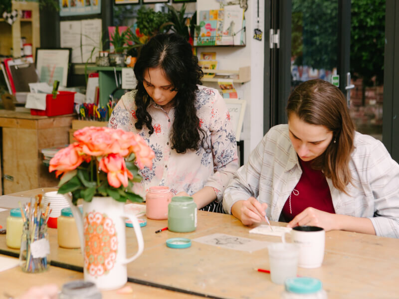 Two women sit at a table, painting ceramic tiles by hand