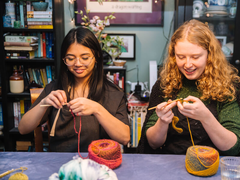 Two women are sitting at a table, crocheting in coloured yarn