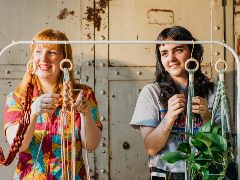 Two women stand at a frame, making macrame planters out of orange and green cord