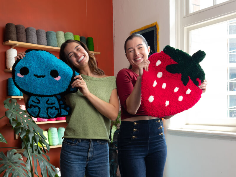 Two women are holding hand tufted rugs and smiling