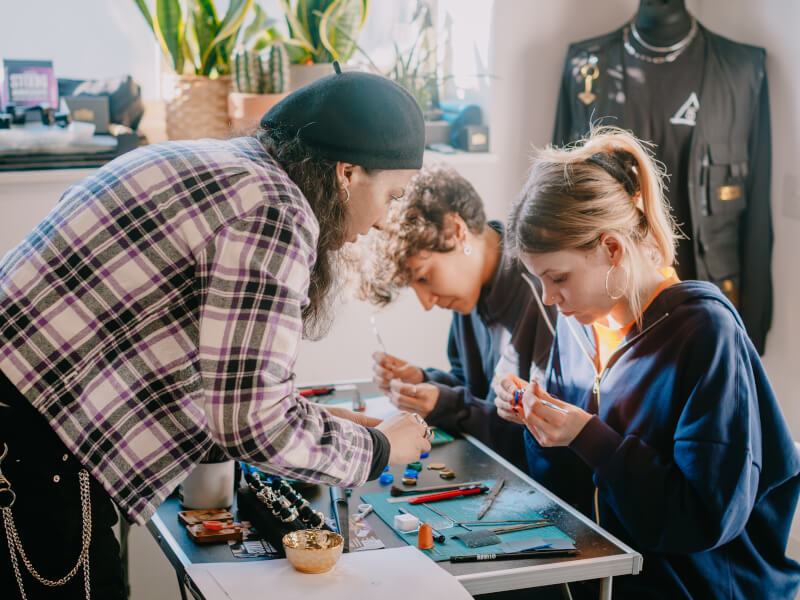 Two women are being taught to make silver rings by a third woman