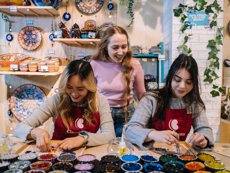 A woman in a pink top is teaching two women sitting in front of her to make mosaic designs
