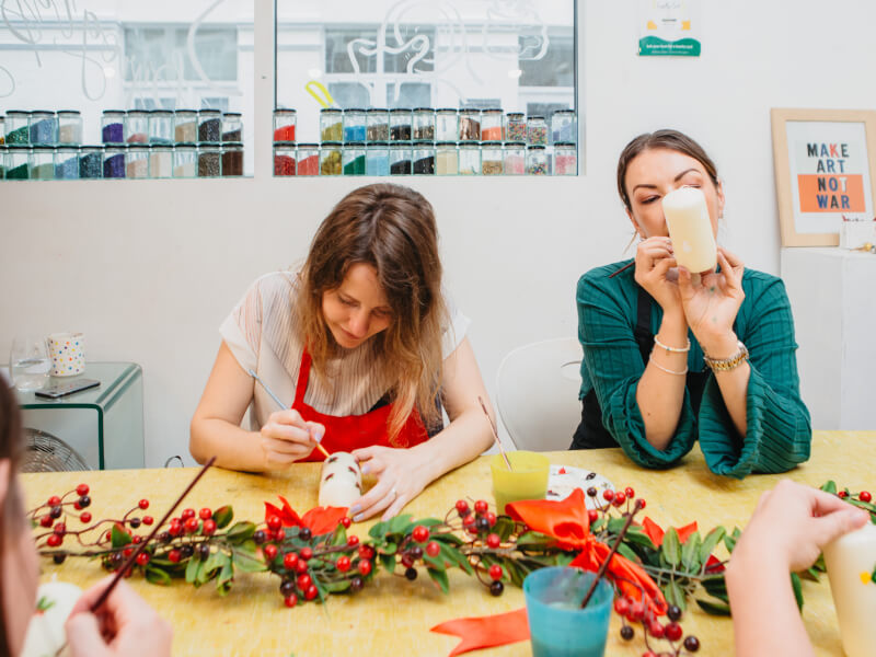 Two women at a candle painting and decorating class are holding pillar candles and painting them with small motifs