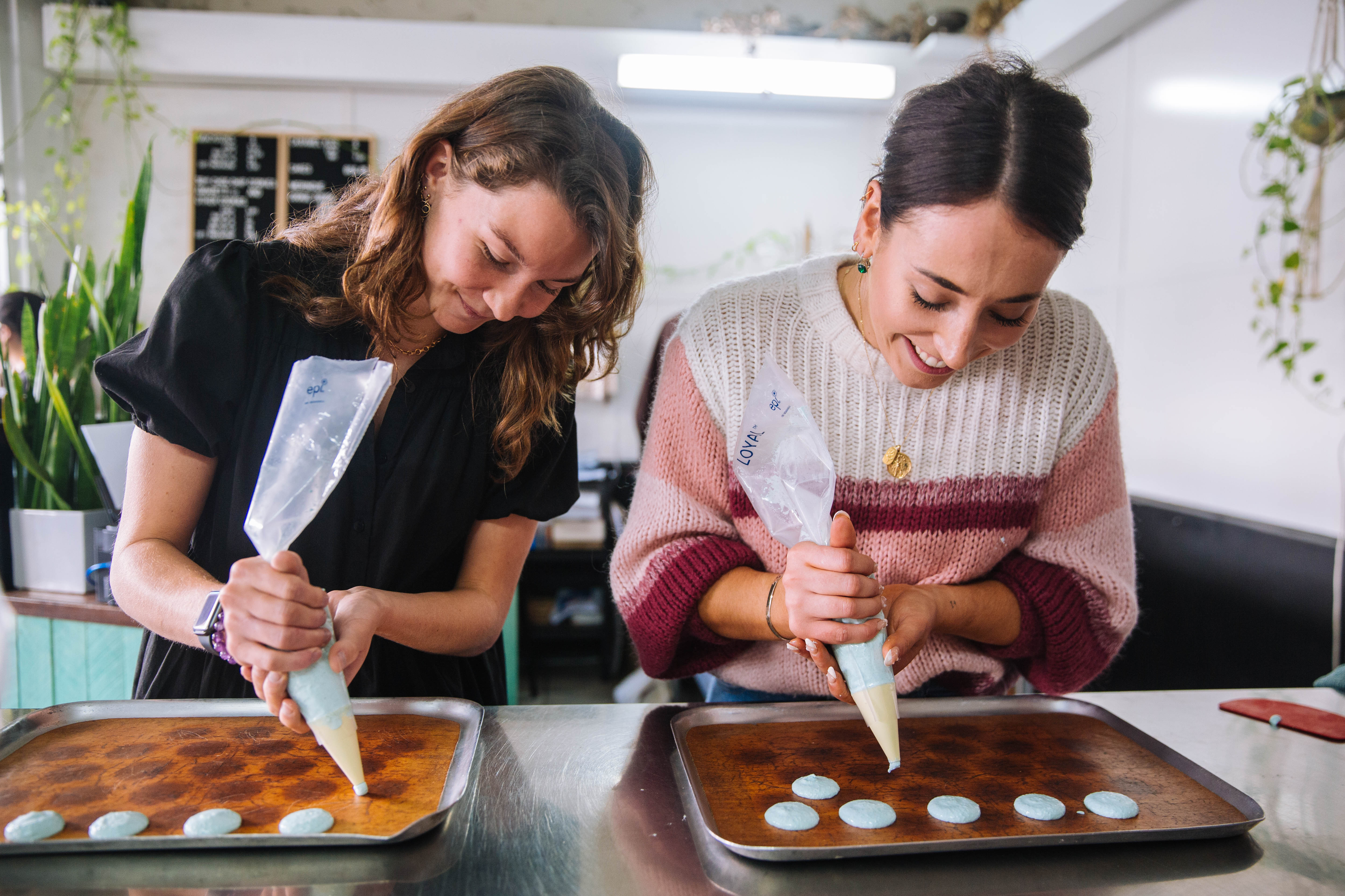 Two women are piping macarons onto a baking tray and smiling