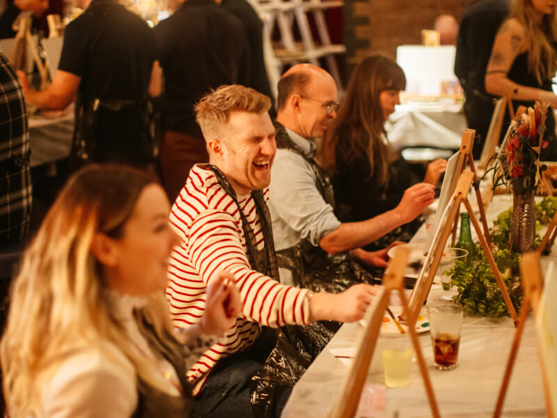 A man is sitting at an easel and laughing at a paint and sip class