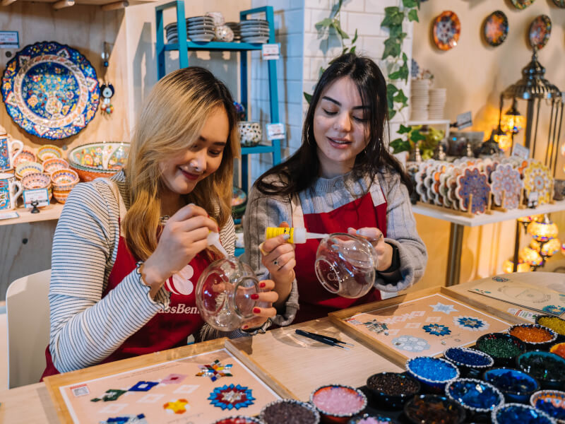 Two women are making mosaic lamps using glass lampshades and coloured tiles