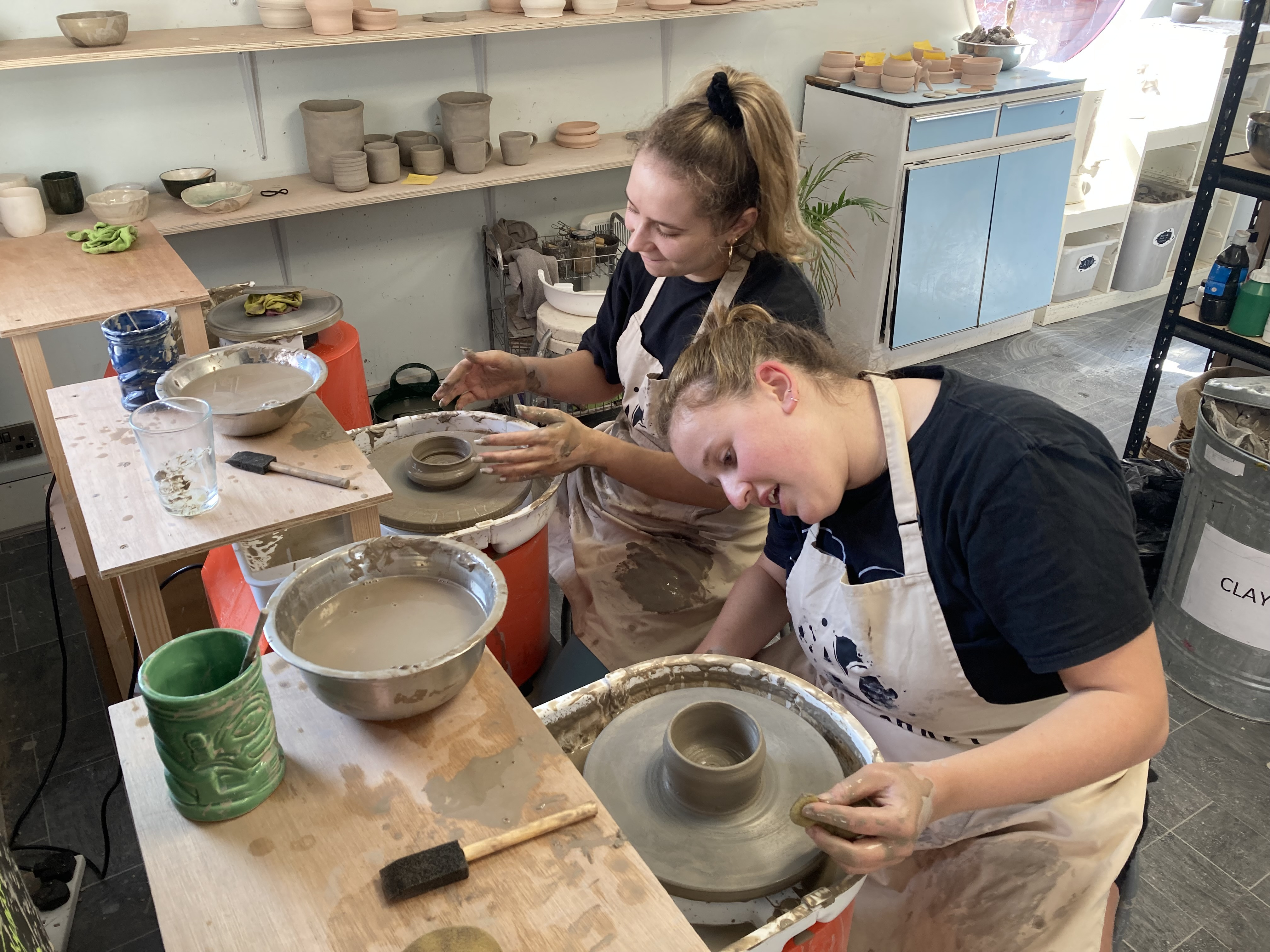 Two laughing women are sitting at pottery wheels, making small round pots