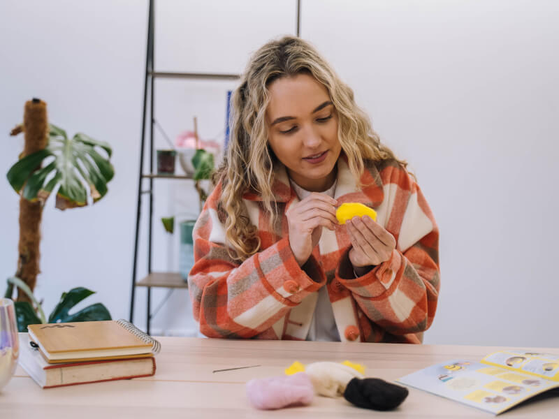 A woman in an orange check jacket is sitting at a table hand sewing yellow felt