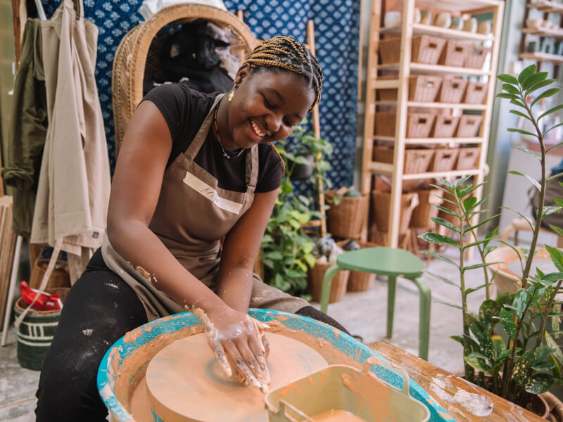 A woman sitting at a pottery wheel is shaping a lump of clay with her hands