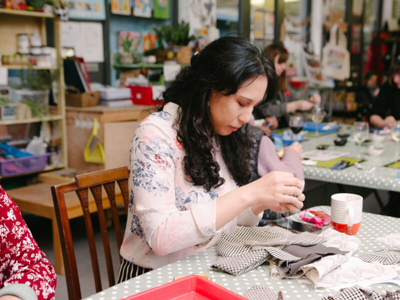 A woman is sitting at a table learning to sew, surrounded by fabric