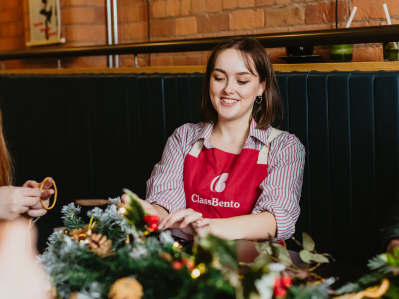 A woman at a wreath making workshop is smiling and arranging greenery