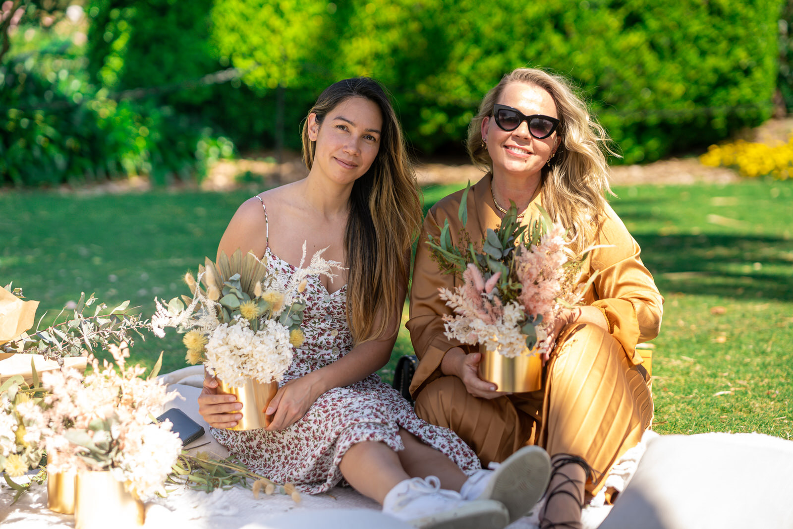 Two women pose, smiling, with their handmade flower arrangements in gold vases