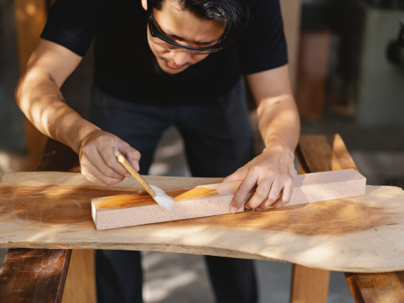 A person is standing at a workbench, applying varnish to a piece of wood at a Manchester woodworking class