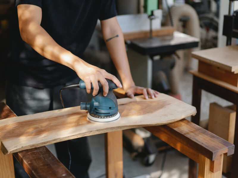 A person is standing at a workbench, sanding a length of wood 