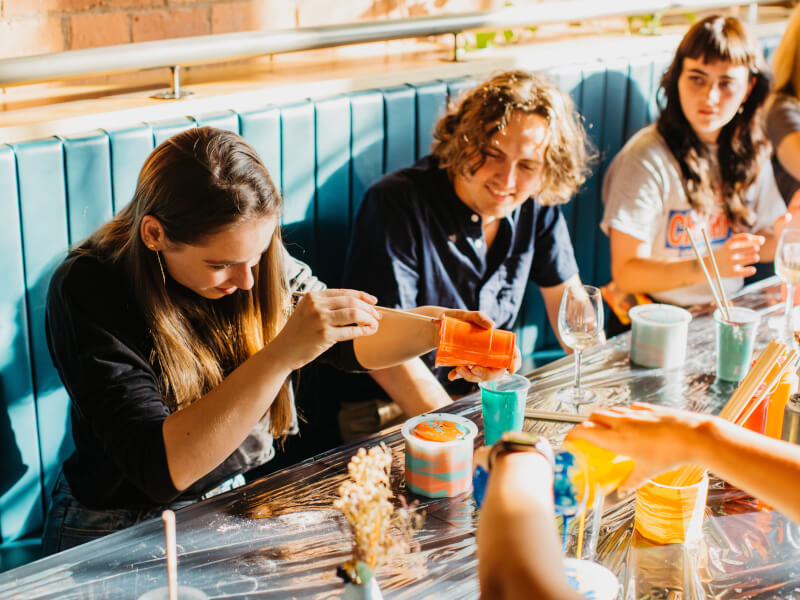 A group of people are sat at a wooden table doing a paint pouring workshop