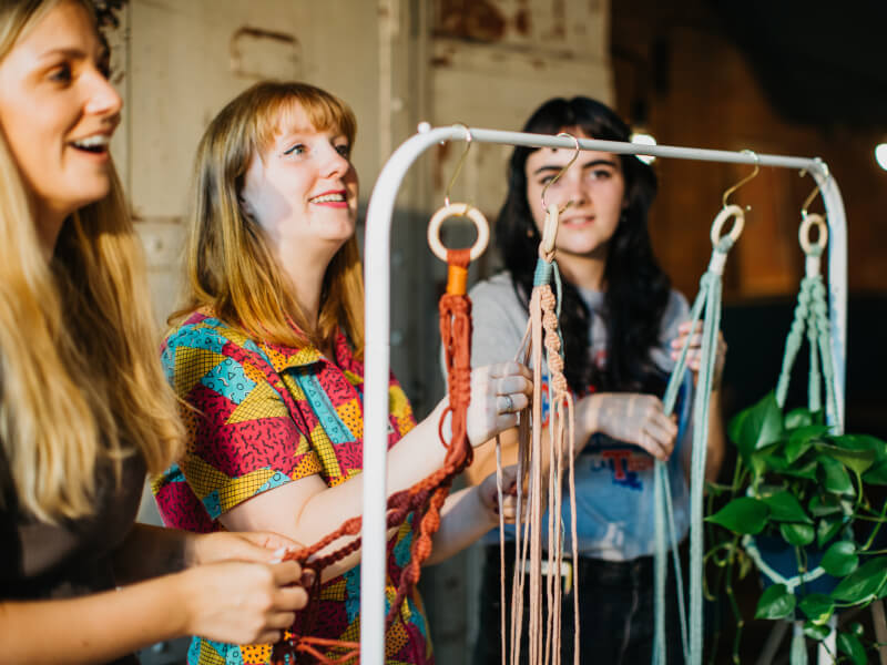 Three laughing women are making macrame plant hangers from brightly coloured cord
