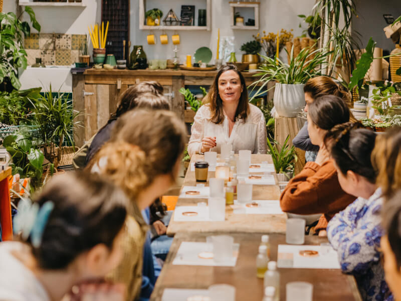 A group of people are sitting at a large wooden table surrounded by plants, making candles