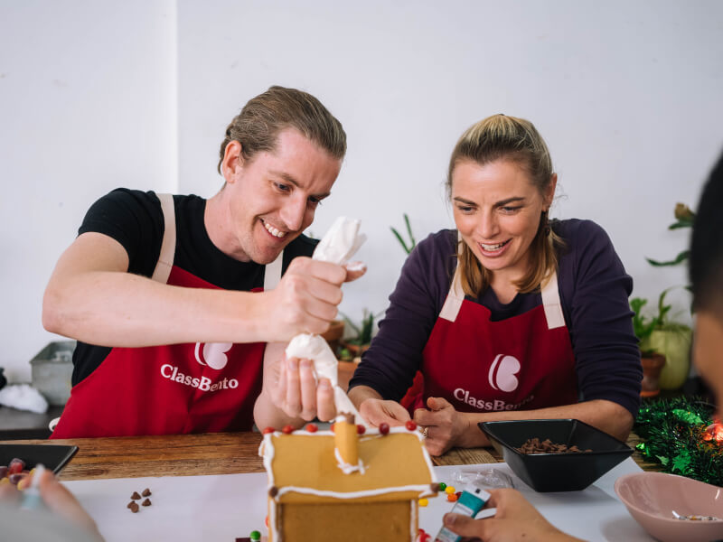 A man and woman sit at a table decorating a gingerbread house with icing